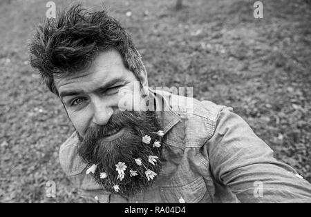 Ragazzo con minor celandine fiori in barba tenendo selfie foto. Hipster con allegro grimace sulla faccia tenendo selfie foto. L'uomo gode di molla, prato verde, sfondo sfocato. Buon umore concetto. Foto Stock