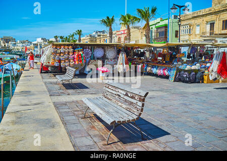 MARSAXLOKK, Malta - 18 giugno 2018: Il lungomare del villaggio di pescatori con panchine presso il porto e il mercato di souvenir sullo sfondo, Giugno 18 Foto Stock