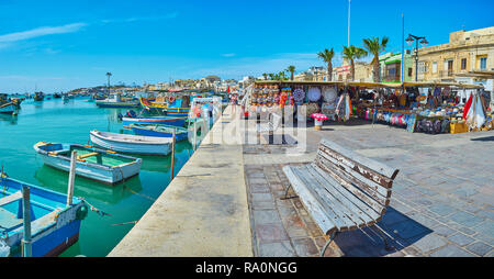 MARSAXLOKK, Malta - 18 giugno 2018: Xatt Is-Sajjieda lungomare con artigianato le bancarelle del mercato e il vecchio legno barche da pesca nella Baia di Marsaxlokk Foto Stock