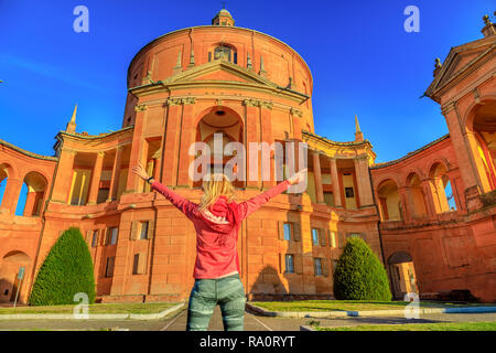Carefree donna con aperto in corrispondenza di bracci a Madonna di San Luca Santuario. Una popolare destinazione turistica di Bologna, Italia. Femmina bionda godendo della storica Basilica alla luce del tramonto. Soleggiato con cielo blu. Foto Stock
