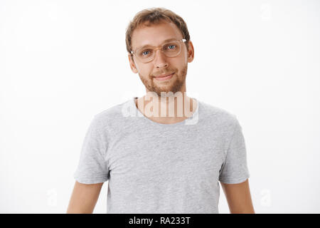 Vita-up shot di carino gentile e bello europeo maschio con la barba in bicchieri e grigio t-shirt sorridente felicemente con rilassato e espressione di sollievo ascoltando la domanda del cliente oltre il muro bianco Foto Stock