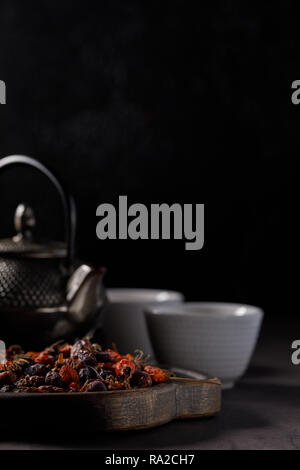 Bollitore con vitamina drink in abbeveratoi e frutti della rosa canina essiccata su una tavola di legno. Il concetto di salute. Ancora vita con tè su un nero backg Foto Stock
