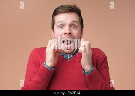 Emozionato sorpreso giovane uomo in maglione rosso mantenere serrati i pugni athis volti, scioccato Foto Stock