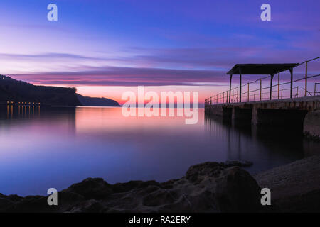 Purpureo tramonto e un molo riflessione su un setoso acqua di mare. Foto Stock