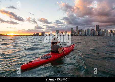 Ragazza avventurosa kayak nella parte anteriore di un moderno centro di Cityscape durante un tramonto spettacolare. Prese a Miami, Florida, Stati Uniti d'America. Foto Stock