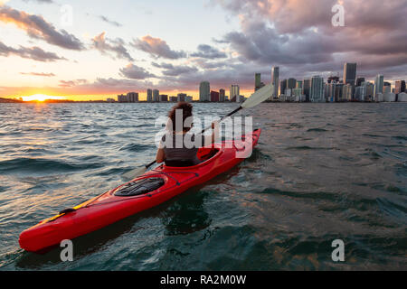 Ragazza avventurosa kayak nella parte anteriore di un moderno centro di Cityscape durante un tramonto spettacolare. Prese a Miami, Florida, Stati Uniti d'America. Foto Stock