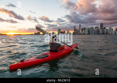 Ragazza avventurosa kayak nella parte anteriore di un moderno centro di Cityscape durante un tramonto spettacolare. Prese a Miami, Florida, Stati Uniti d'America. Foto Stock