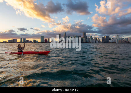 Ragazza avventurosa kayak nella parte anteriore di un moderno centro di Cityscape durante un tramonto spettacolare. Prese a Miami, Florida, Stati Uniti d'America. Foto Stock