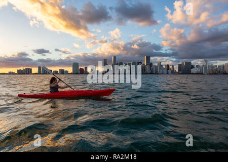 Ragazza avventurosa kayak nella parte anteriore di un moderno centro di Cityscape durante un tramonto spettacolare. Prese a Miami, Florida, Stati Uniti d'America. Foto Stock