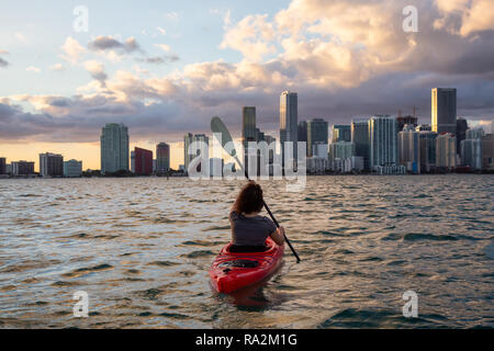 Ragazza avventurosa kayak nella parte anteriore di un moderno centro di Cityscape durante un tramonto spettacolare. Prese a Miami, Florida, Stati Uniti d'America. Foto Stock