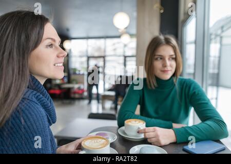 Sorridente due giovani donne in una caffetteria con caffè arte e amaretti in piastre, ragazze parlando, stagione invernale indossando maglioni caldi. Foto Stock