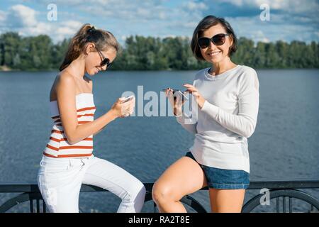 Mamma e figlia passeggiate in estate il parco vicino al fiume, utilizzando i telefoni cellulari, parlando e sorridente. Foto Stock