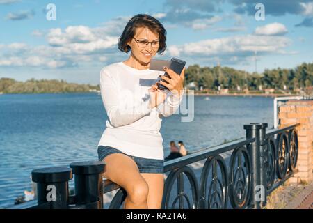 Donna di mezza età con telefono mobile e business card. Femmina dello smartphone utilizza guardando la scheda Foto Stock