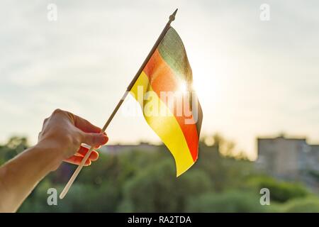 Mano che tiene la Germania bandiera una finestra aperta. Lo sfondo è blu cielo, la silhouette della città, il tramonto. Foto Stock
