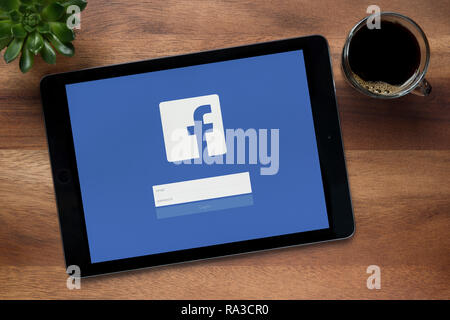 Facebook è visto su un tablet iPad, su di un tavolo di legno lungo con un caffè espresso e una pianta di casa (solo uso editoriale). Foto Stock