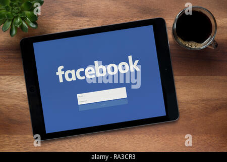 Facebook è visto su un tablet iPad, su di un tavolo di legno lungo con un caffè espresso e una pianta di casa (solo uso editoriale). Foto Stock