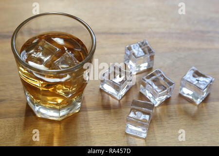 Un piccolo bicchiere di whiskey è mostrato accanto a cubetti di ghiaccio su di una superficie di legno, con retroilluminazione naturale. Foto Stock