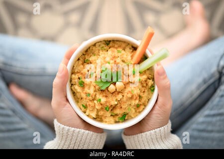 A piedi nudi donna tenendo un recipiente con hummus, la carota e il sedano seduta sul pavimento Foto Stock