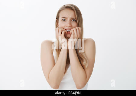 Vita-up shot di inorridito nel panico giovani precari e attraente donna con capelli biondi e carino moli di mordere le unghie e accigliato occhi popping in telecamera spaventato e timido oltre il muro grigio Foto Stock