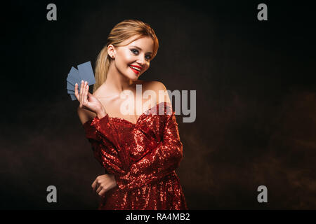 Sorridente ragazza attraente in rosso vestito lucido azienda carte da gioco isolato su nero Foto Stock