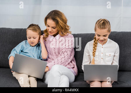 Bella donna con figlio e figlia seduti sul divano e utilizzando i notebook in appartamento Foto Stock