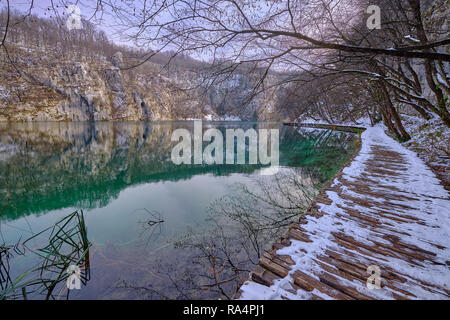 A piedi in legno sentiero coperto di neve a fianco di color verde lago. Sul lato scogliera si riflette nell'acqua. Scena nel Parco Nazionale di Plitvice Foto Stock