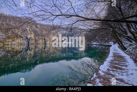 A piedi in legno sentiero coperto di neve a fianco di color verde lago. Sul lato scogliera si riflette nell'acqua. Scena nel Parco Nazionale di Plitvice Foto Stock