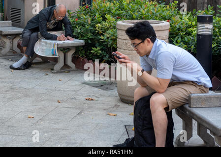 Vecchio e nuovo: le differenze generazionali di Hong Kong dove un uomo vecchio scansiona il suo giornale, matita in mano, mentre un giovane uomo scansiona il suo telefono cellulare. Foto Stock