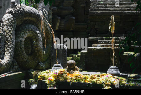 Bali tradizionale tempio con il nero di sculture in pietra e le offerte per il dio fatto da foglie di banano Foto Stock