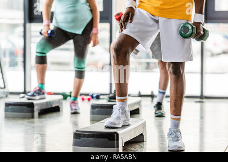 Immagine ritagliata di African American sportsman e i suoi amici esercizio con manubri su piattaforme di passo in palestra Foto Stock