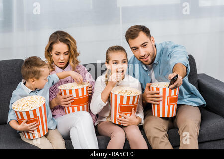 Padre sorridente seduto sul divano e modifica dei canali mediante il dispositivo di controllo remoto con azienda familiare striped benne e mangiare popcorn in appartamento Foto Stock
