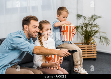 Padre sorridente seduto sul divano e modifica dei canali mediante il dispositivo di controllo remoto con bambini holding striped secchielli per popcorn in appartamento Foto Stock