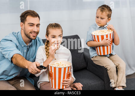 Padre sorridente seduto sul divano e modifica dei canali mediante il dispositivo di controllo remoto con bambini azienda benne a strisce e mangiare popcorn in appartamento Foto Stock