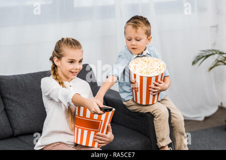 Sorridente ragazza seduta sul divano e cambiare i canali con telecomando con il fratello mangiare popcorn in appartamento Foto Stock