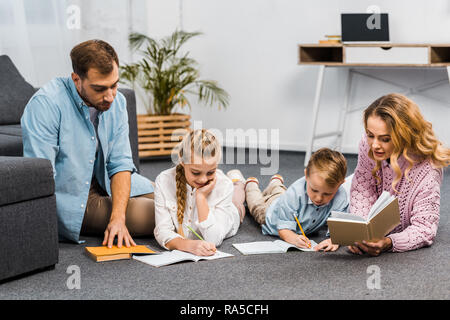 I genitori la lettura di libri mentre i fratelli iscritto nei libri di esercizio sul pavimento in appartamento Foto Stock