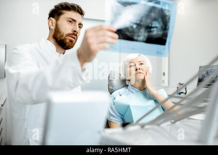 Preoccupato elder donna durante la consultazione con bel dentista che mostra vedute x-ray nello studio dentistico Foto Stock