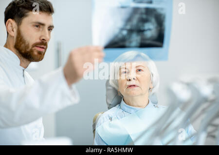 Preoccupato elder donna durante la consultazione con bel dentista che mostra vedute x-ray nello studio dentistico Foto Stock