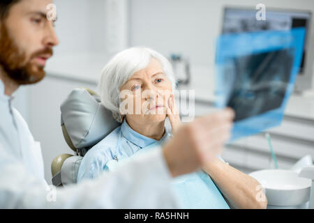 Preoccupato elder donna durante la consultazione con bel dentista che mostra vedute x-ray nello studio dentistico Foto Stock