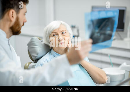 Preoccupato elder donna durante la consultazione con bel dentista che mostra vedute x-ray nello studio dentistico Foto Stock