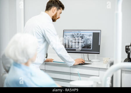 Dentista che mostra le riprese panoramiche della ganascia sul computer per l'anziana signora durante la consultazione in studio dentistico Foto Stock