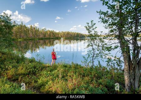 Donna di mezza età in piedi presso il lago, foresta, gli alberi che riflettono, Hossa National Park, Ruhtinansa, Suomussalmi, Kainuu, Finlandia Foto Stock