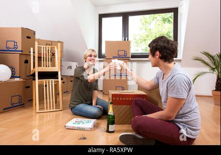 Coppia giovane seduto in una stanza vuota in un nuovo appartamento e sollevando i loro bicchieri di champagne per festeggiare in movimento insieme Foto Stock