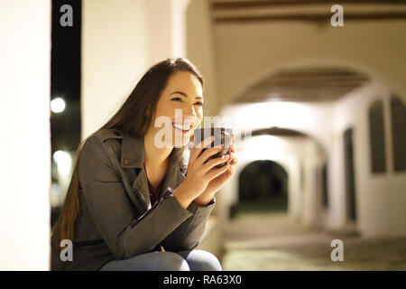 Donna felice di bere il caffè guardando lontano seduto in una strada della città di notte Foto Stock