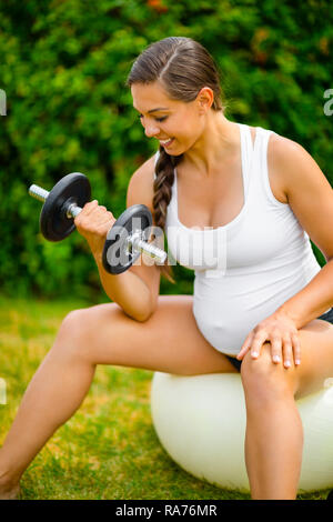 Donna incinta sorridere mentre il sollevamento dei pesi sulla palla ginnica Foto Stock