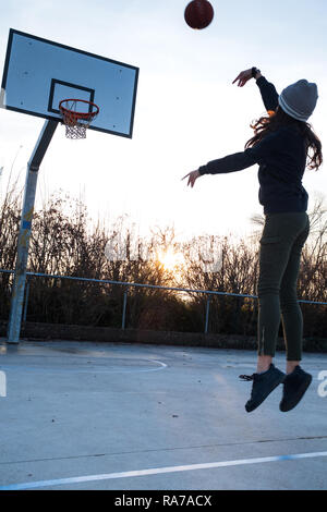 Young Teen girl, metà aria, tiro jump shot sul campo di pallacanestro, retroilluminato al tramonto, composizione verticale, spazio di copia Foto Stock