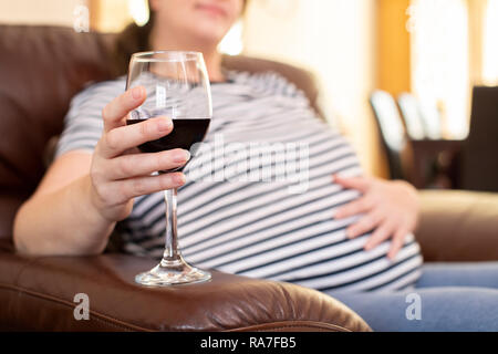 Close up della donna incinta di bere vino rosso seduto sul divano di casa Foto Stock