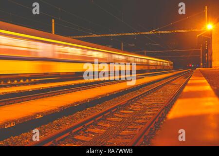 Bianco e blu treno passa una piccola scoperchiata stazione ferroviaria di notte nella Repubblica Ceca. Sfocata treno di movimento Foto Stock