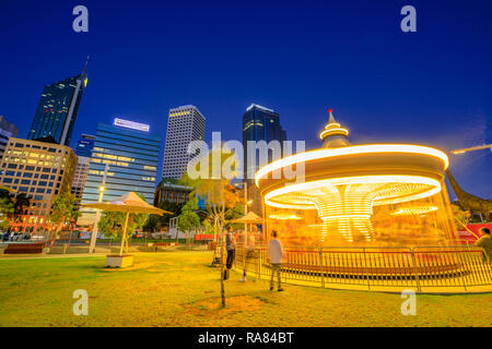 Perth, Western Australia - Jan 6, 2018: veneziana tradizionale giostra illuminata di notte gira veloce a Elizabeth Quay. Esplanade con grattacieli del quartiere degli affari e sullo sfondo. Scena notturna. Foto Stock