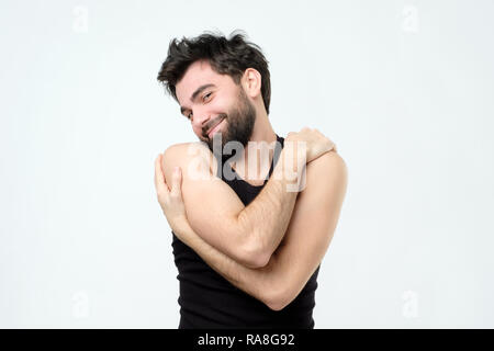 Persone, auto amore e narcisismo concetto. Gioiosa bel giovane ispanico uomo sorridente con piacere mentre abbracciando stesso, tenere le braccia intorno al suo sh Foto Stock