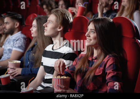 Vista dal lato della bella dai capelli scuri ragazza in maglietta rossa godendo la visione di film interessanti al cinema. Bella femmina spettatore seduto vicino al ragazzo biondo e mangiare popcorn dal rosso coppa. Foto Stock